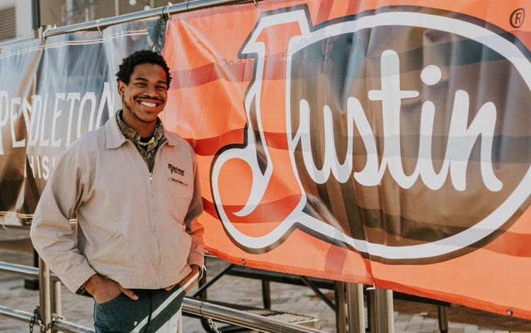 Man stands next to a fence with Justin Boots Banner on the fence.